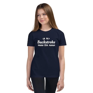 Peut inclure: Un t-shirt bleu marine avec l'inscription blanche "IN MY Backstroke ERA" et des vagues. Le t-shirt est de style col rond.