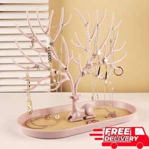 May include: A pink deer-shaped jewellery organiser with antler-like branches for hanging necklaces. The base is an oval tray. Various necklaces and rings are displayed. The image includes the text "FREE DELIVERY".