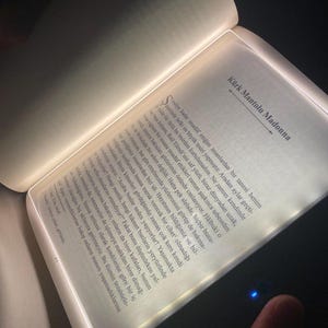Può includere: Un libro aperto con una luce LED integrata, che illumina le pagine. Il testo è in lingua straniera, con il titolo "Kürk Mantolu Madonna" visibile. Il libro è su una superficie scura.