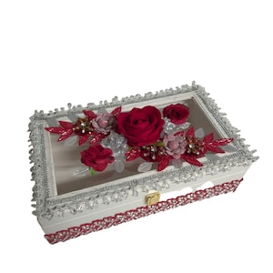 May include: A white rectangular box with a clear top, decorated with red roses and floral arrangements. The box is trimmed with silver and red lace, and has a gold-coloured clasp.