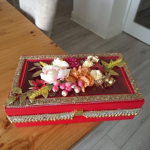 May include: A red decorative box with a clear lid, decorated with artificial flowers in pink, peach, and white. The box is trimmed with gold and silver detailing and a gold clasp. The box is likely used for gifting.