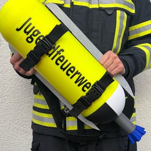 Könnte beinhalten: Ein leuchtend gelber zylindrischer Tank mit schwarzer Aufschrift Jugendfeuerwehr, ausgestattet mit schwarzen verstellbaren Gurten, einer weiß-schwarzen Basis und einer blauen Düse am Ende. Der Tank wird horizontal gehalten und hat ein graues Verbindungsstück.