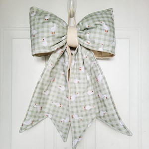 May include: A large decorative bow made of green and white checkered fabric, adorned with white geese with red feet and beaks. The bow has a beige center and is hanging on a white door. The bow is a charming addition to farmhouse decor.