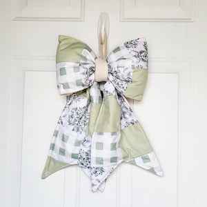 May include: A large, decorative bow with a patchwork design, featuring shades of green, white, and floral patterns. The bow is made of fabric and has a cream-colored band in the center. It is hanging on a white door.