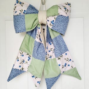 May include: A large decorative bow made of quilted fabric featuring a patchwork design. The pattern consists of alternating squares of solid sage green, blue floral print, and a white background with a blue blueberry and green leaf print.