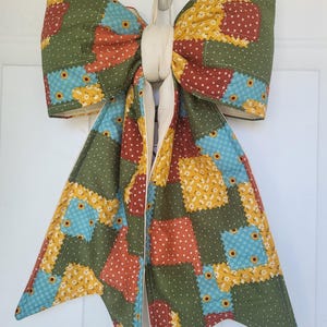 May include: A large, decorative bow made of patchwork fabric, featuring squares of green, yellow, blue, and red with floral and polka dot patterns. The bow is suspended by a cream-colored loop, suitable for door or wall decor.