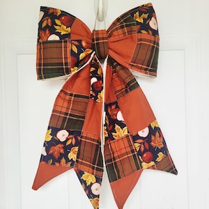 May include: A large fabric bow featuring a patchwork design of dark navy blue fabric with a print of red apples and yellow maple leaves, alternating with solid burnt orange fabric and a brown, green, and orange plaid fabric pattern.