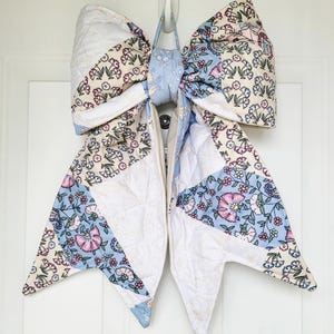 May include: A large, quilted bow in shades of white, light blue, and pink, with floral patterns. The bow is hanging on a white door, and is likely a decorative item for a door or wall.