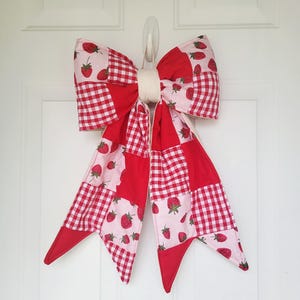May include: A large, decorative bow with a patchwork design, featuring red gingham, strawberry-printed fabric, and solid red sections. The bow is cream-colored in the center and is designed to be hung on a door. The bow is a festive decoration.