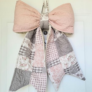May include: A large, quilted bow in soft pink and patchwork patterns. The bow features a quilted design and is suspended by a fabric loop. The patchwork includes floral, gingham, and geometric patterns in pink, gray, and white.