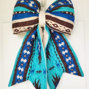 May include: A large, decorative bow made of quilted fabric. The bow features a Southwestern-inspired pattern in shades of turquoise, brown, blue, and cream. The bow is attached to a white loop, suggesting it's designed for hanging as a door or wall decoration.