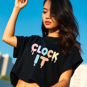 Puede incluir: Top corto negro con el texto "CLOCK IT" en colores pastel. Las letras tienen un efecto de goteo. La modelo está al aire libre con un cielo azul de fondo.