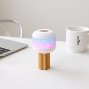 Puede incluir: Una pequeña lámpara decorativa con una parte superior blanca texturizada y una base cilíndrica acanalada de color dorado. La parte superior emite un degradado de luz azul, rosa y morada. En el fondo se ve una taza de café blanca con la palabra "coffee".