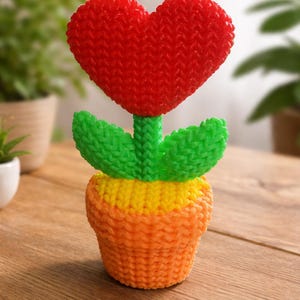 Puede incluir: Una escultura de flores tejida con una flor roja en forma de corazón, hojas verdes y una maceta amarilla y naranja. La decoración hecha a mano se coloca sobre una superficie de madera, con un fondo borroso de plantas verdes. La flor mide unos 15 cm de alto.