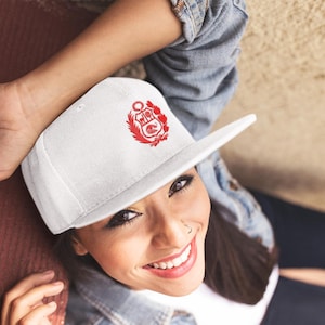 May include: A white baseball cap featuring a red emblem design. The cap has a flat brim and a structured crown. The emblem appears to be a crest or coat of arms, with intricate details. The cap is a classic style, suitable for casual wear.