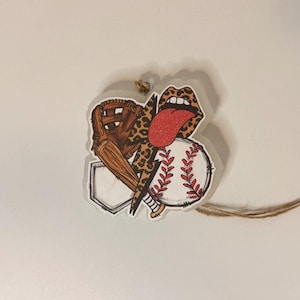 May include: A baseball-themed ornament featuring a baseball, glove, and bat. The design includes a leopard print lightning bolt and lips with a red tongue. The ornament is attached to a twine string.