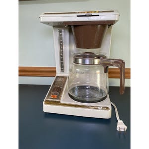 May include: A vintage Norelco 12-cup coffee maker. The appliance is white with brown accents, including the filter basket and handle. A clear glass carafe sits below. The coffee maker has a dial and a water level indicator.