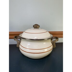 Może przedstawiać: Kremowy garnek emaliowany z pokrywką i dwoma uchwytami. Garnek ma brązowe paski i srebrną obręcz. Pokrywka zwieńczona jest mosiężnym pokrętłem. Uchwyty są również mosiężne. Garnek jest prawdopodobnie w stylu vintage.