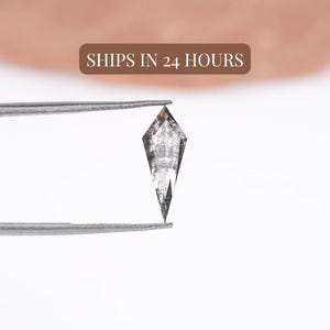 May include: A kite-shaped salt and pepper diamond with a translucent, speckled grey and black appearance held by metal tweezers. The gemstone features a pointed top and bottom with faceted edges, displaying natural internal inclusions throughout.