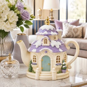 May include: A ceramic teapot shaped like a cottage with a cream-colored base, a light blue door, and a purple roof covered in white snow. The teapot features a gold finial on top, a curved handle, a spout, and small green potted plants by the door.
