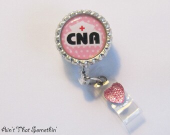 May include: A silver badge holder with a pink and white polka dot design featuring a white nurse's cap with a red cross and the text "CNA". A pink heart charm is attached to the badge holder.