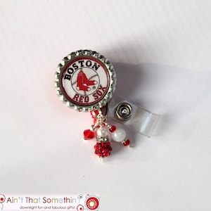May include: A Boston Red Sox themed badge reel. The reel features a bottle cap design with the team logo and colors. It is adorned with red and white beads and a clear plastic clip.