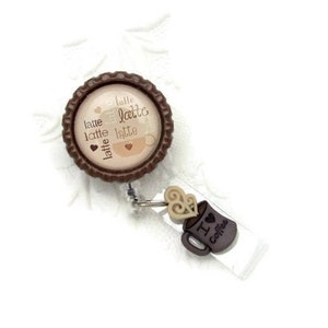 May include: Brown and beige retractable badge holder with a coffee cup design and the text "latte latte latte latte" on the front. The holder has a brown plastic clip and a clear plastic strap.