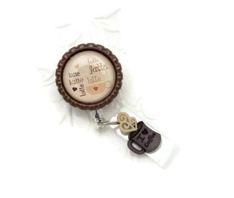 May include: Brown and beige retractable badge holder with a coffee cup design and the text "latte latte latte latte" on the front. The holder has a brown plastic clip and a clear plastic strap.