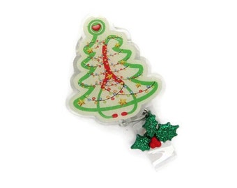 May include: A Christmas-themed badge reel featuring a green Christmas tree design with a red ribbon and string lights. The badge reel also has a green glitter holly and red berry accent. The badge reel is on a white background.