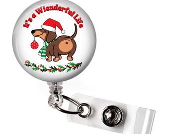May include: A white retractable badge reel with a Christmas-themed design. The design features a cartoon dachshund wearing a Santa hat and scarf, holding an ornament, with the text "It's a Wienderful Life".