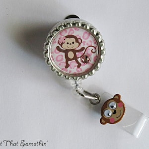 May include: A pink and brown monkey with a flower on its head is on a silver and white retractable badge holder. The badge holder has a clear plastic strap and a small monkey charm.