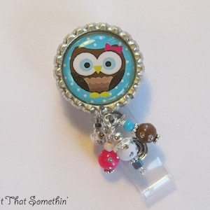 May include: A silver badge holder with a blue and white owl design. The owl has a pink bow and large eyes. The badge holder has a clear plastic strap and a variety of colorful beads.