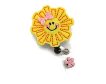 May include: A yellow felt sun with a pink bow and a smiling face attached to a retractable badge holder with a pink flower charm.
