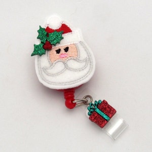 May include: A red and white Santa Claus face with a green holly sprig on top, attached to a red retractable badge holder with a green and red glitter gift charm.