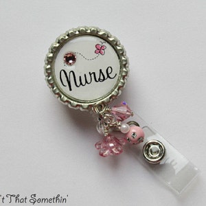 May include: A silver and white badge holder with a pink butterfly and the word "Nurse" on the front. The badge holder has a clear plastic retractable reel and pink and white beads.
