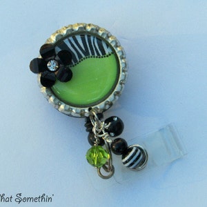 May include: A green, black, and white zebra print bottle cap badge reel with a black flower and rhinestones. The reel is attached to a clear plastic badge holder.
