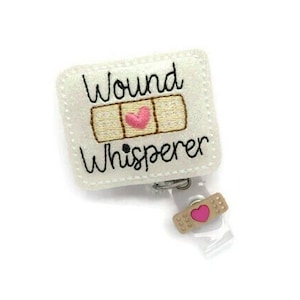May include: A white felt badge reel with the words "Wound Whisperer" embroidered in black. The design includes a tan bandage with a pink heart in the center. A small bandage charm with a pink heart is attached.