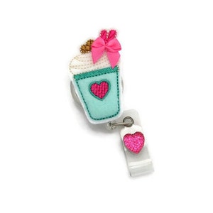 May include: A teal blue and white cupcake with a pink bow and a pink heart on the side, attached to a retractable badge holder with a pink glitter heart.