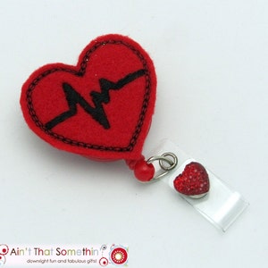 May include: Red felt heart with a black embroidered EKG line. The heart is attached to a clear plastic badge holder with a red rhinestone heart charm.