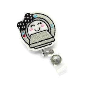 May include: White embroidered badge reel featuring a cartoon CT scan with a black and white bow and a smiling face. The reel has a silver clip and a clear plastic strap.