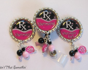 May include: Three circular bottle cap badge reels featuring black and pink designs with the text Rx Pharmacy, Rx Pharmacist, and Rx Pharmacy Tech. Each piece is decorated with dangling black and pink beads and silver-toned metal accents.