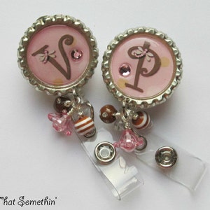 May include: Two pink bottle cap charms with the letters "V" and "P" in brown, decorated with rhinestones and small brown and white beads. The charms are attached to a clear plastic retractable badge holder.