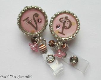 May include: Two pink bottle cap charms with the letters "V" and "P" in brown, decorated with rhinestones and small brown and white beads. The charms are attached to a clear plastic retractable badge holder.