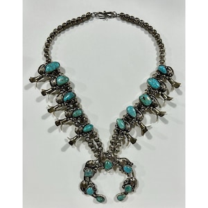 May include: A squash blossom necklace featuring polished turquoise stones set in silver. The piece includes a central naja pendant, multiple silver beads, and silver flower-shaped blossoms along the sides, all connected by a silver hook clasp.