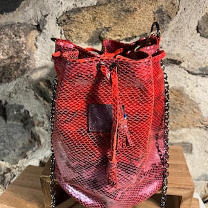 May include: Red bucket bag with a chain strap. The bag features a drawstring closure and a small leather tag. The exterior has a snakeskin pattern. A small tassel hangs from the drawstring. The bag is sitting on a wooden surface.