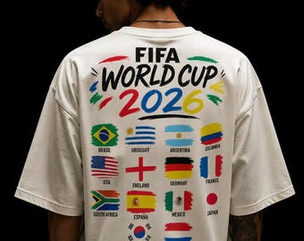 May include: An off-white t-shirt featuring the text FIFA WORLD CUP 2026 in black, red, and yellow brushstroke lettering. Below the text are twelve national flags arranged in a grid, each with its country name printed in black underneath.