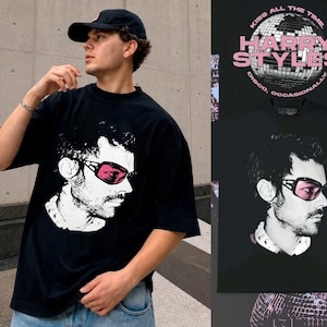 Puede incluir: Una camiseta negra de manga corta con un gráfico blanco de una persona con gafas de sol de cristales rosas. La parte posterior de la camiseta muestra un gráfico de bola de discoteca con el texto "KISS ALL THE TIME HARRY STYLES DISCO OCCASIONALLY".