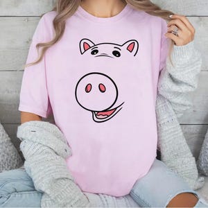 以下が含まれることがあります： 淡いピンク色の半袖Tシャツで、黒とピンクの豚の顔のグラフィックプリントが施されています。デザインには上部に2つの耳、中央に大きな鼻、下部に舌が見える笑顔の口が描かれています。