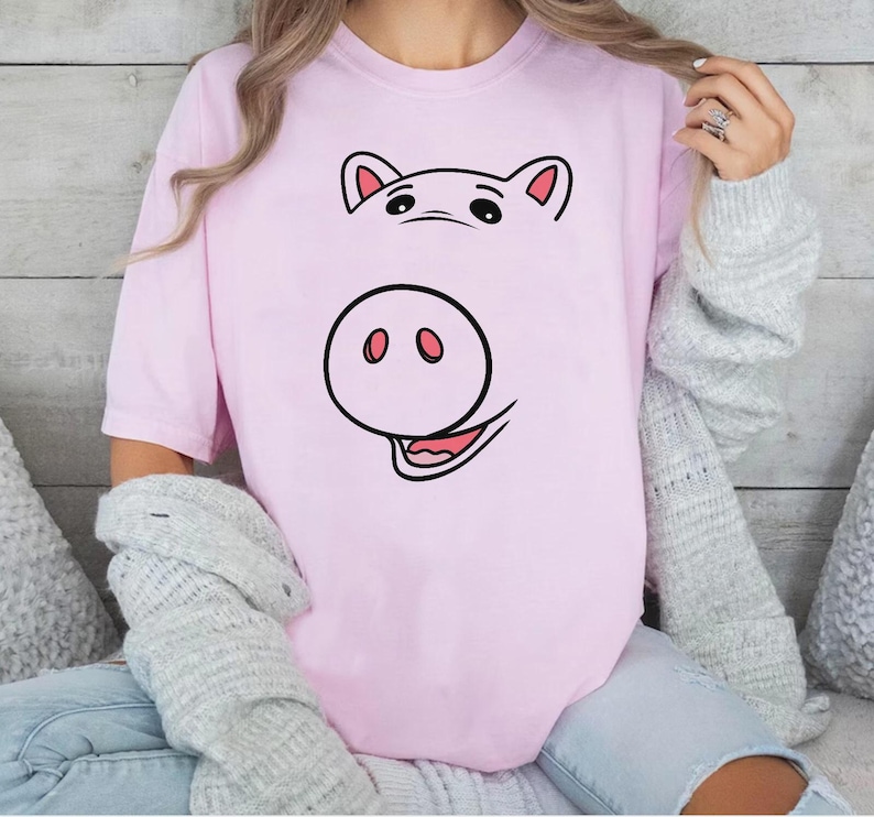 以下が含まれることがあります： 淡いピンク色の半袖Tシャツで、黒とピンクの豚の顔のグラフィックプリントが施されています。デザインには上部に2つの耳、中央に大きな鼻、下部に舌が見える笑顔の口が描かれています。