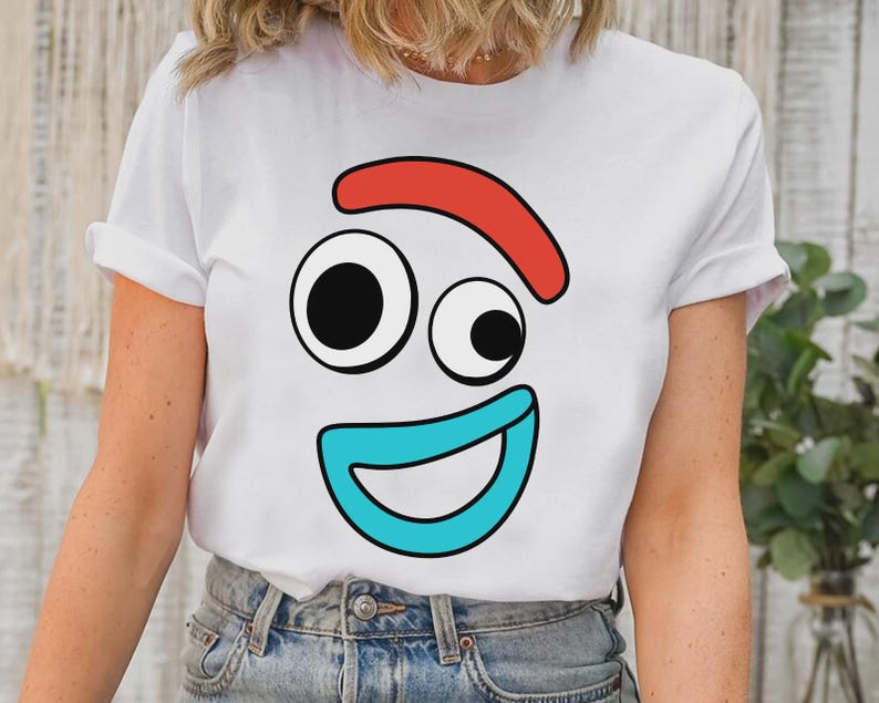 以下が含まれることがあります： 白い半袖Tシャツで、漫画の顔のグラフィックプリントが施されています。デザインには、赤い曲線の眉毛、左右非対称の黒と白の丸い目、そして胸の中央に配置された幅の広い水色の曲線の口が含まれています。